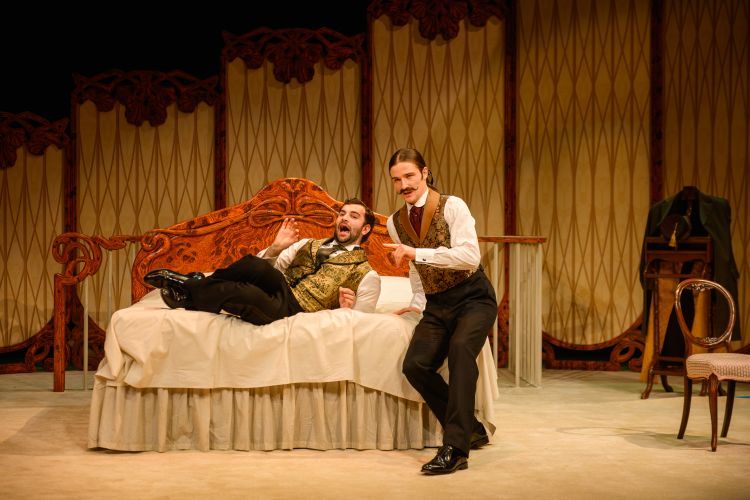 Two male performers in flamboyant regency style dress including gold patterned waistcoats are singing out to the audience. One is lying back on a double bed with ornate wooden headboard and the other is perched on the corner of the bed. Panelled screens are in the background descending in height